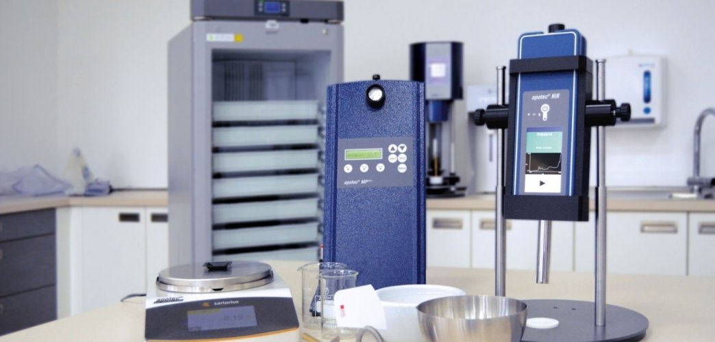 apotec Apothekenmaschinen stehen an einem Tisch mit einer Waage, einem Mörser, und hinten steht ein Kühlschrank von apotec.