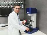 Prozess Ein Apotheker hält eine Kruke in der einen Hand und drückt mit der anderen Hand auf ein Display vor ihm, auf einer apotec Maschine
